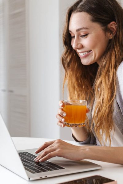 Pretty cheerful young woman having breakfast while sitting at the kitchen table, working on laptop computer, drinking juice